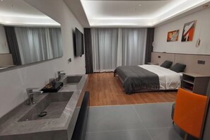 A modern hotel room with a large bed, a built-in desk, a large mirror, a vanity with a sink, and a shower.