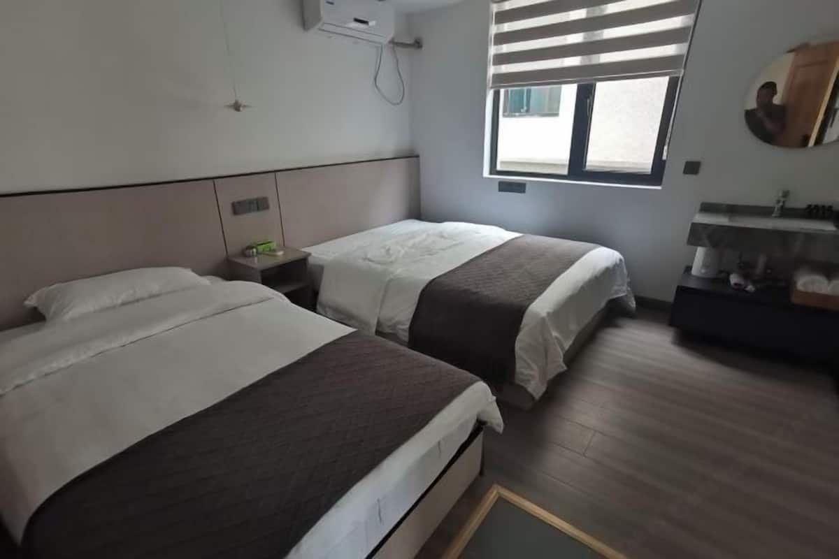 A hotel room with two beds, a window with blinds, a bedside table with a lamp, and a small mirror on the wall.