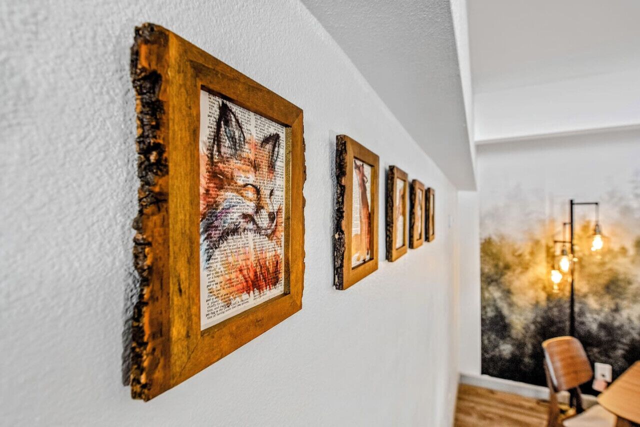 A wall with a series of framed artworks, including a fox painting, in a room with a wooden floor and a window with a view of trees.