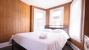 A neatly made bed with white linens and rolled towels, a wooden paneled wall, and a window with blinds.
