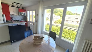 A compact kitchen with a red cabinet, a microwave, and a gas stove. There is a dining table with a woven basket and a chair. The room has large windows with a view of a residential area.