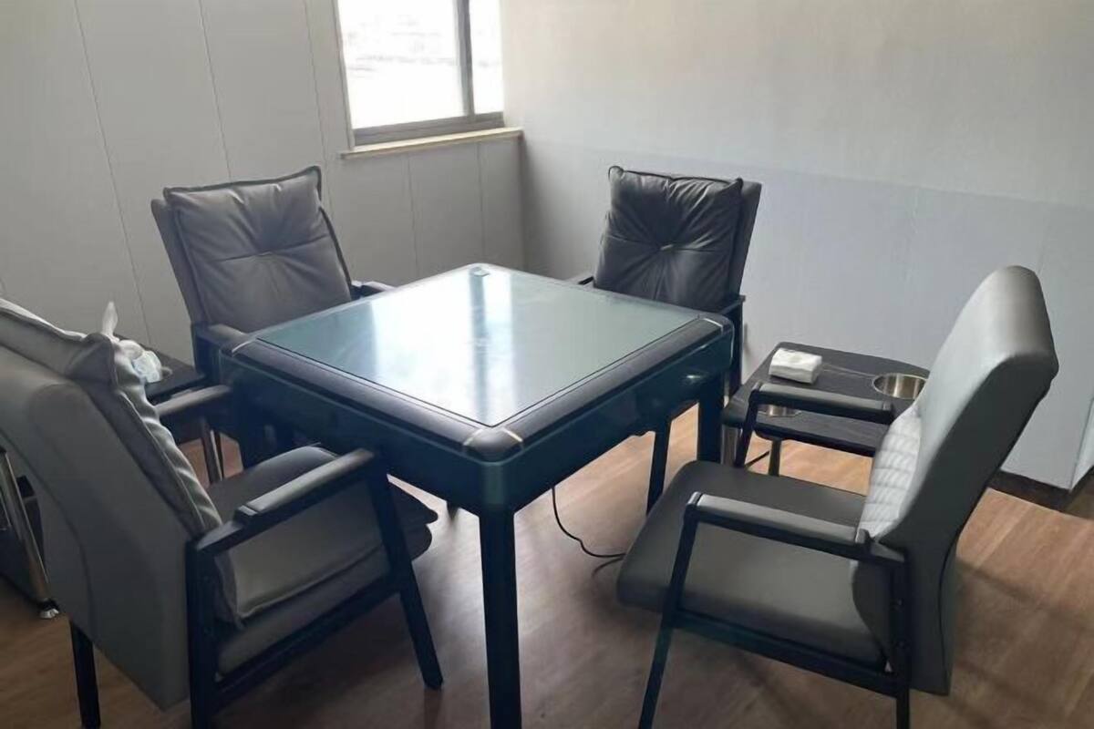 A room with a glass-top table surrounded by four chairs, a small table with a kettle, and a window providing natural light.