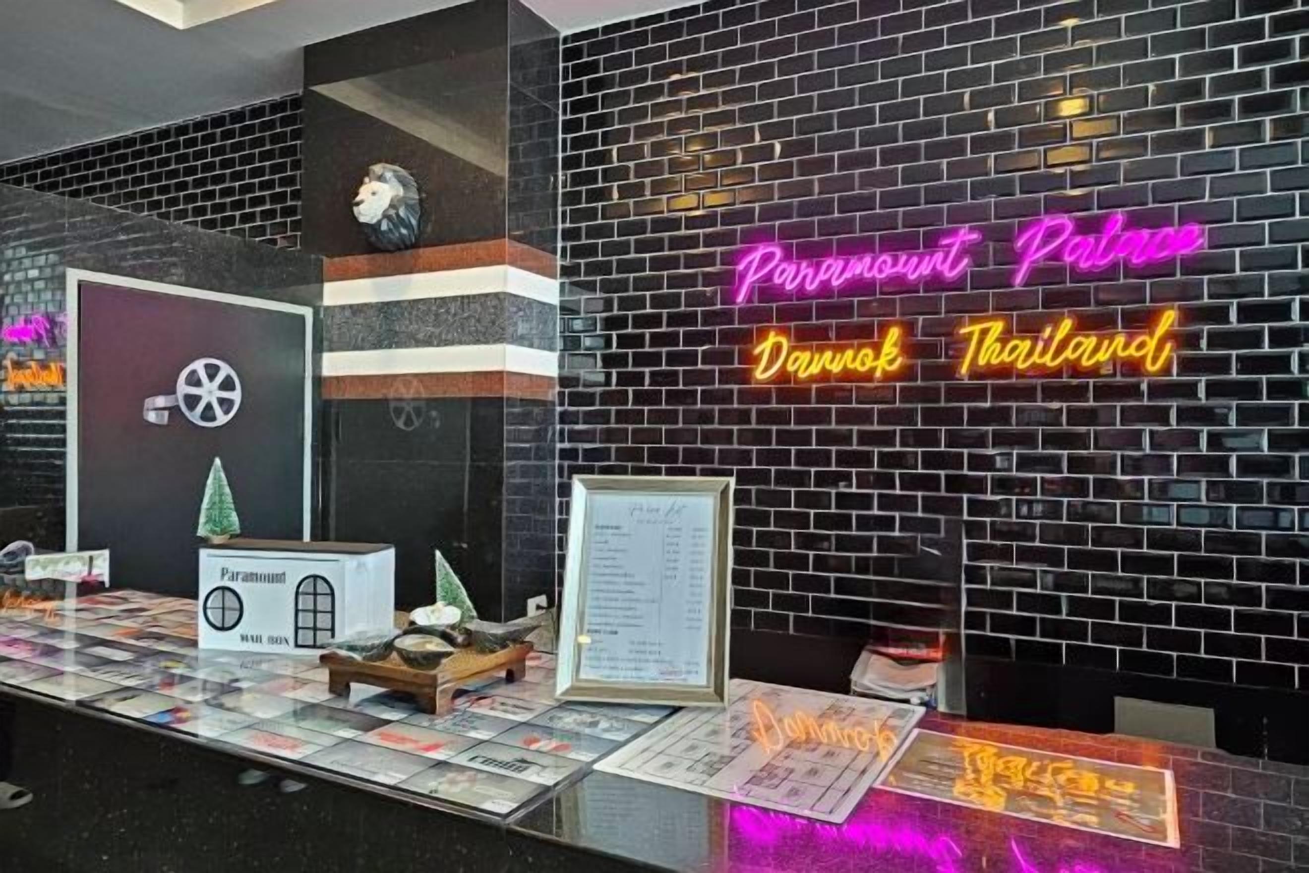 A hotel reception area with a neon sign for 'Paramount Palace Danub Thailand,' a reception desk, and a menu board.