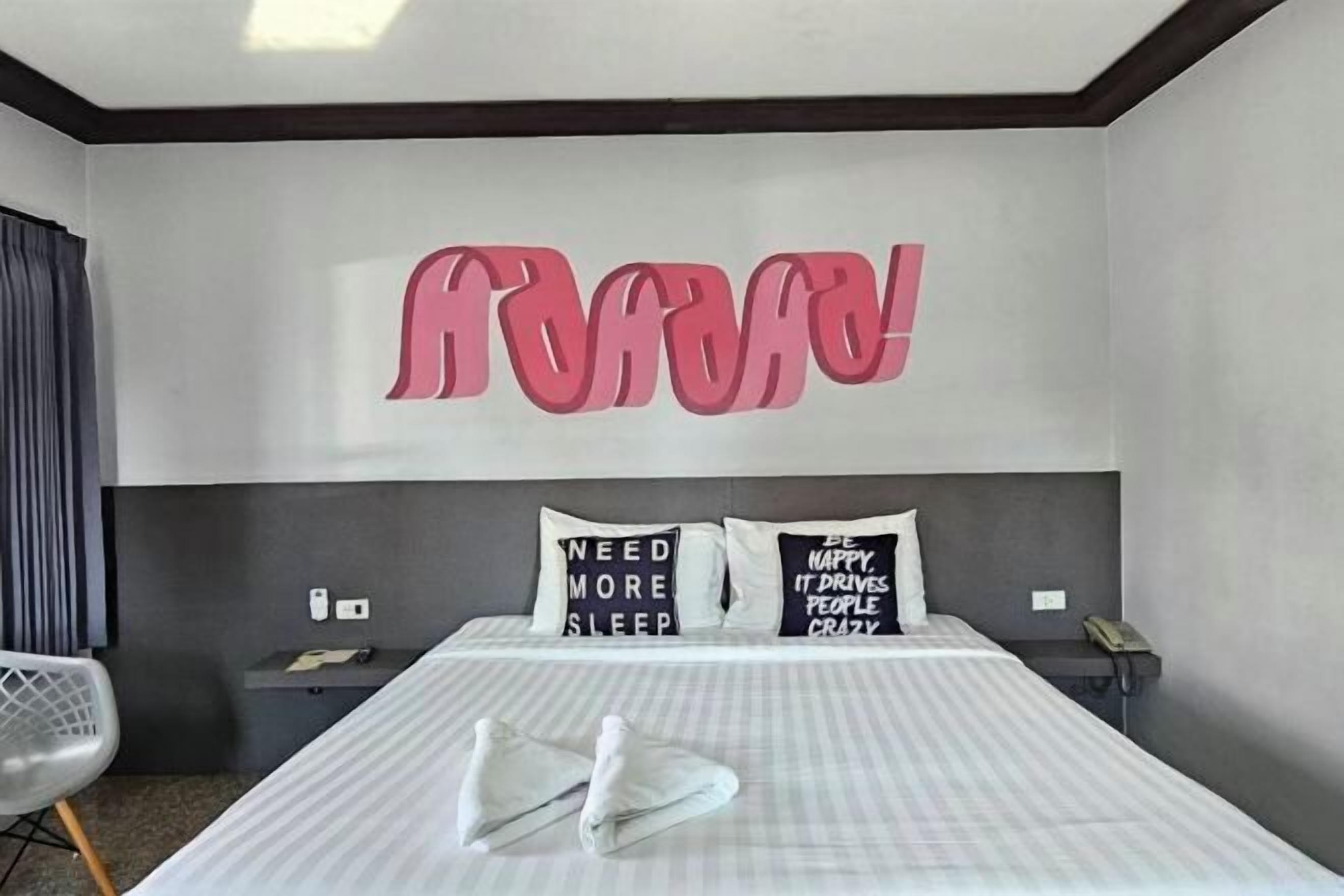 Een moderne hotelkamer met een groot bed, twee kussens met motiverende citaten, een grijze stoel en een muur met grote roze tekst.