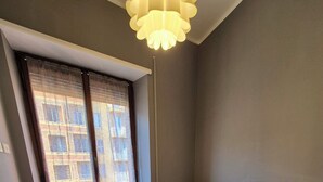 A small, modern living room with a grey sofa, a white coffee table, and a small white table. There is a window with blinds, a ceiling light, and a radiator. - Appartamento al Centro di Roma Esquilino Colosseum Vicino Metro (Roma)
