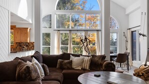 Een moderne woonkamer met een ruime hoekbank, een ronde salontafel en uitzicht op herfstbomen door grote ramen.