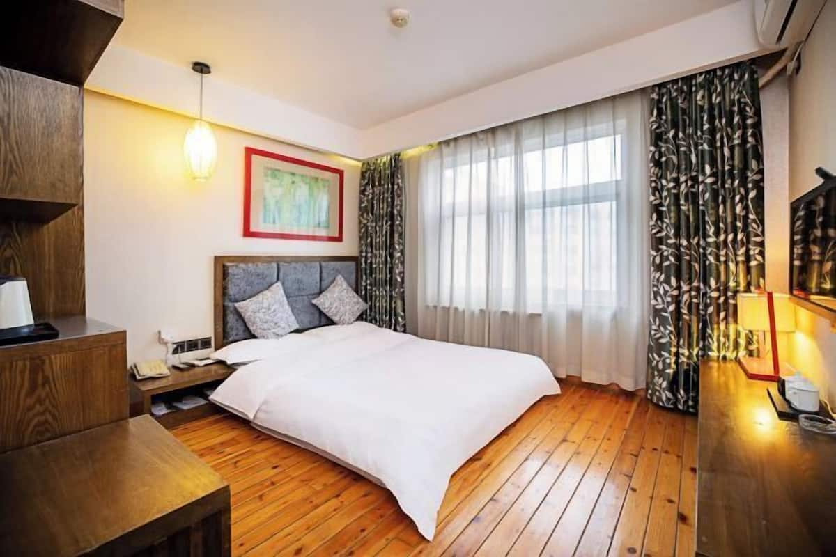 Een hotelkamer met een houten vloer, een bed met een witte dekbedovertrek, een nachtkastje, een lamp en een raam met dunne gordijnen.