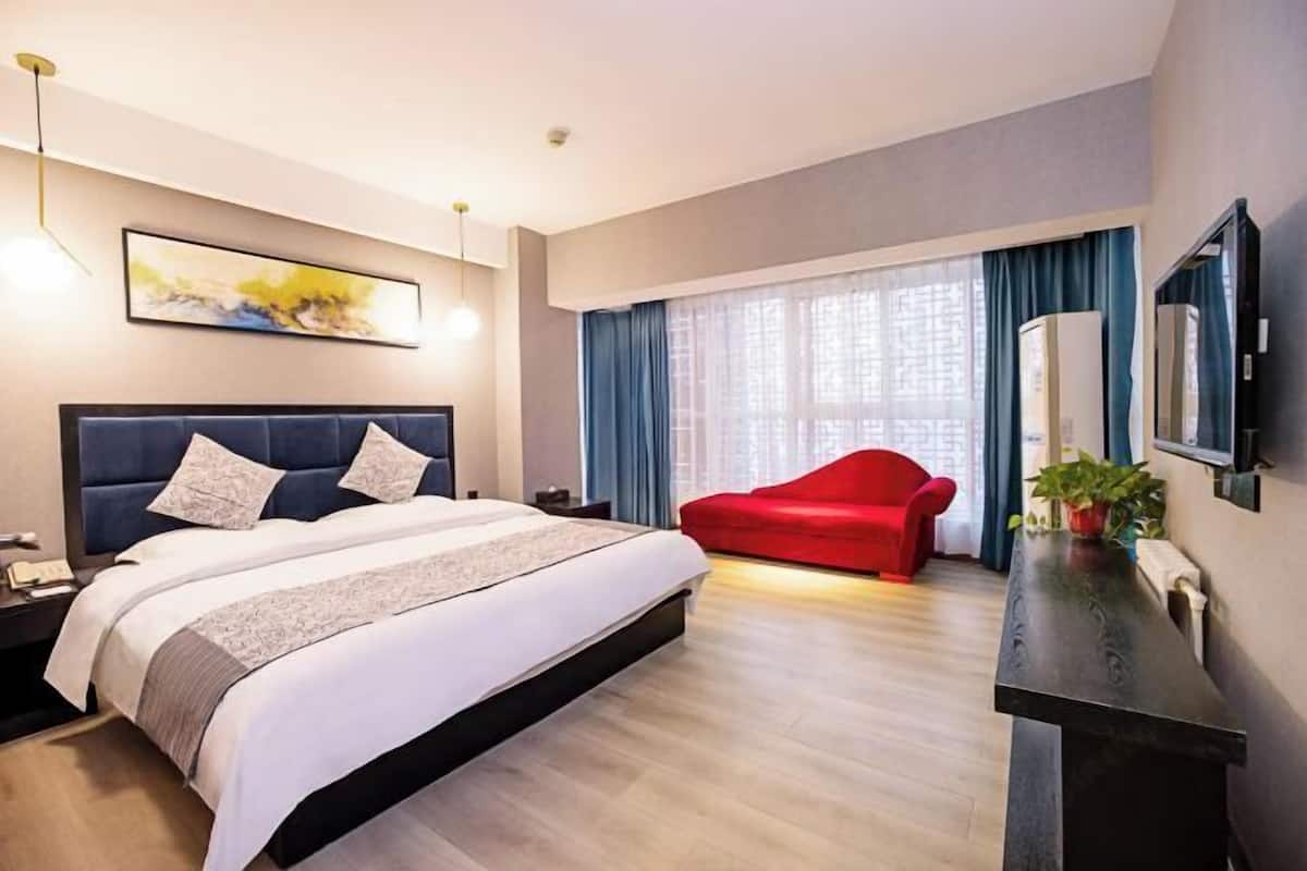 Een moderne hotelkamer met een groot bed, een rode bank, een bureau met een plant en een groot raam met gordijnen.
