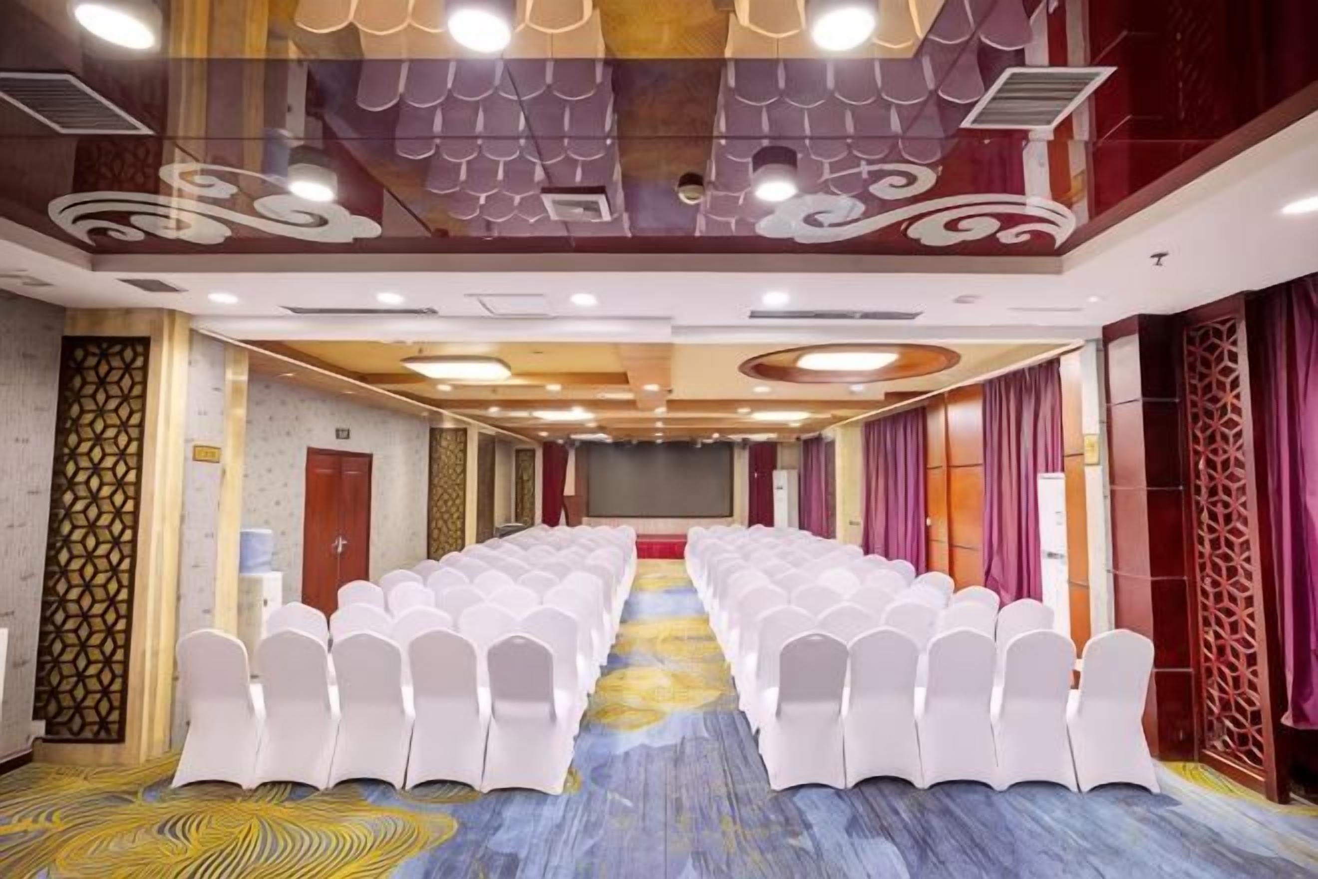Une salle de conférence avec des rangées de chaises blanches, une scène et un tapis à motifs.