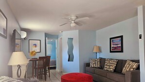 A living room with a grey sofa, a red ottoman, a dining table with chairs, and a ceiling fan. - The Snapper, at Paradise Cove (Englewood)