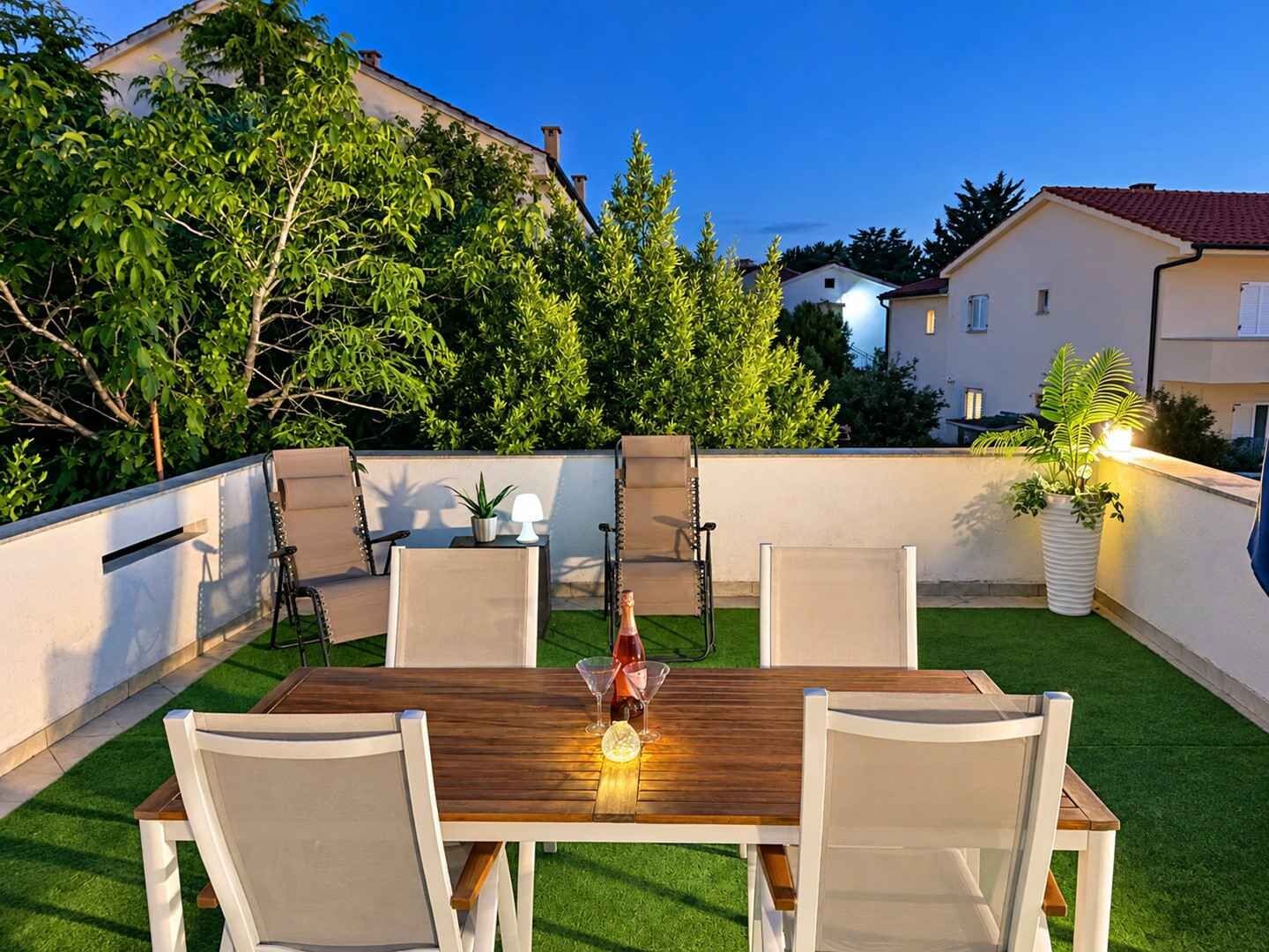 Een terras buiten met een houten tafel en vier stoelen, omringd door een lage muur en groen.