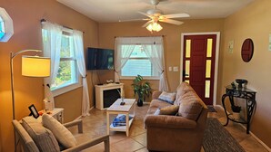 Un salon comprenant un canapé, un fauteuil, un téléviseur, une cheminée, un ventilateur de plafond et une porte rouge.