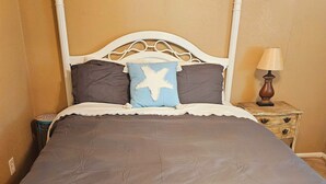 Un lit avec une tête de lit en métal blanche, une couvre-lit marron foncé et un oreiller bleu orné d’une étoile blanche. Au-dessus du lit, on peut voir deux tableaux encadrés accrochés au mur, une table de chevet avec une lampe et une petite table sur laquelle est posé un objet décoratif.