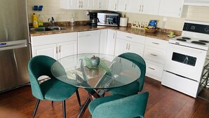 A modern kitchen with white cabinets, a stainless steel refrigerator, a microwave, an oven, and a glass dining table with two teal chairs. - Cozy one bedroom apartment  (Peterborough)