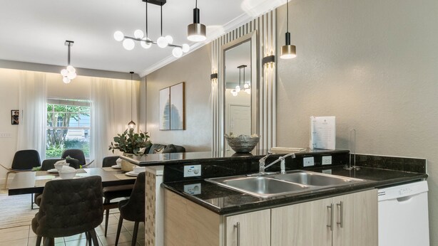 A modern kitchen with a central island, black countertops, and stainless steel appliances. There is a dining area with a table and chairs, and a large window providing natural light. - Last Minute Disney Getaway! Townhouse with Private Pool. Book Now! #65617 (Orlando)
