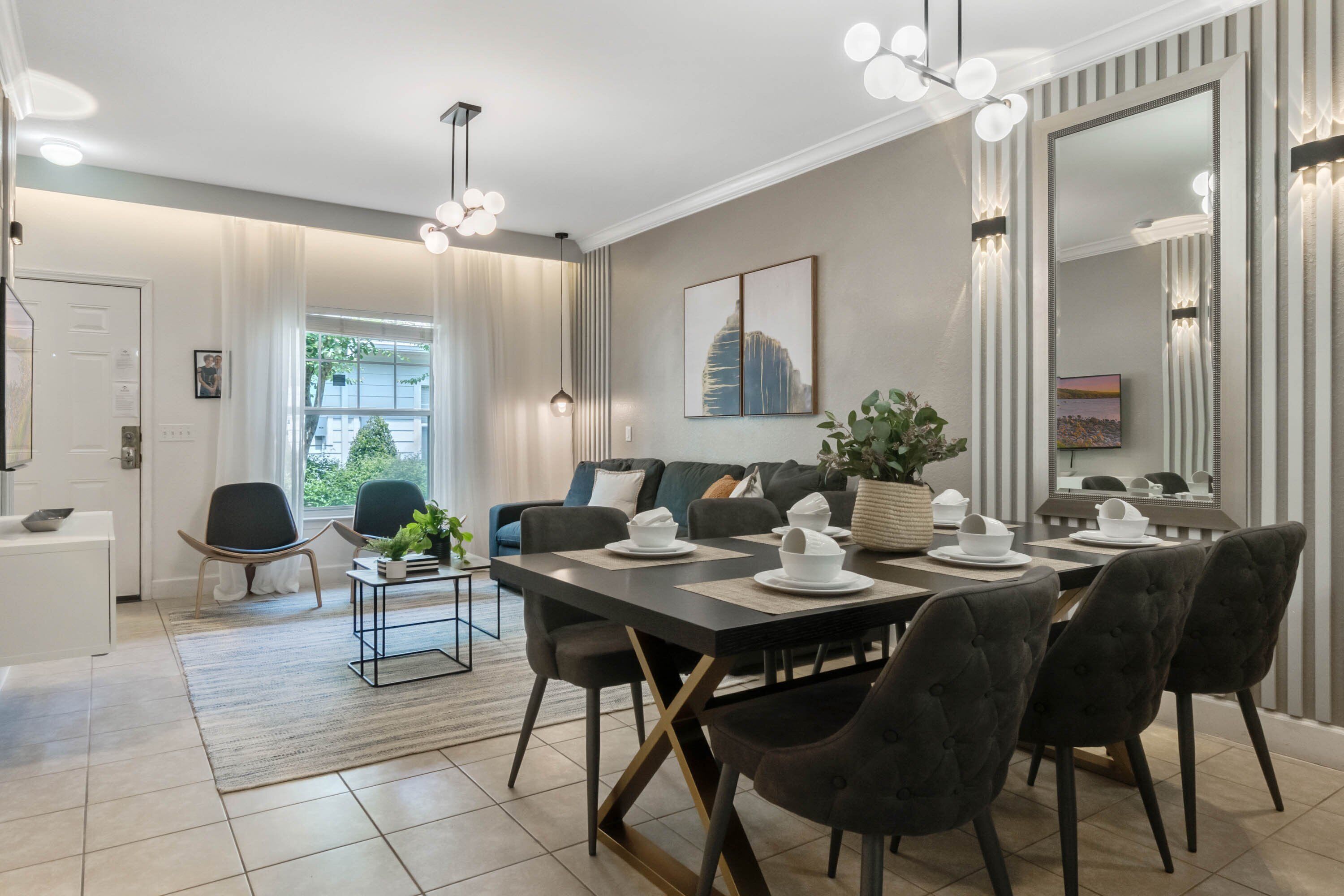 A modern dining area with a long table set for four, surrounded by dark upholstered chairs. A large mirror and a framed cityscape artwork adorn the walls.