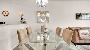 Un espace repas moderne avec une table en verre, des chaises beiges, un lustre, une horloge et un canapé en arrière-plan.
