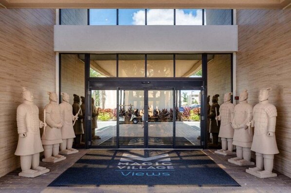 A modern building entrance with glass doors, a blue mat with "Magic Village Views" text, and stone statues on either side.