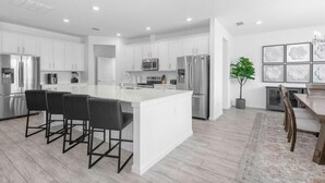 A modern kitchen with white cabinetry, stainless steel appliances, a central island with bar stools, and a dining area with a wooden table and chairs. - Last Minute Disney Getaway! Villa with Private Pool & Hot Tub. Book Now! #116945 (Orlando)