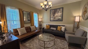 A living room with a brown leather sofa, a grey armchair, a round glass coffee table, and a chandelier.