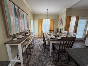 A dining area with a wooden table set for four, surrounded by chairs. There is a rug on the floor, a ceiling light fixture, and a painting on the wall.