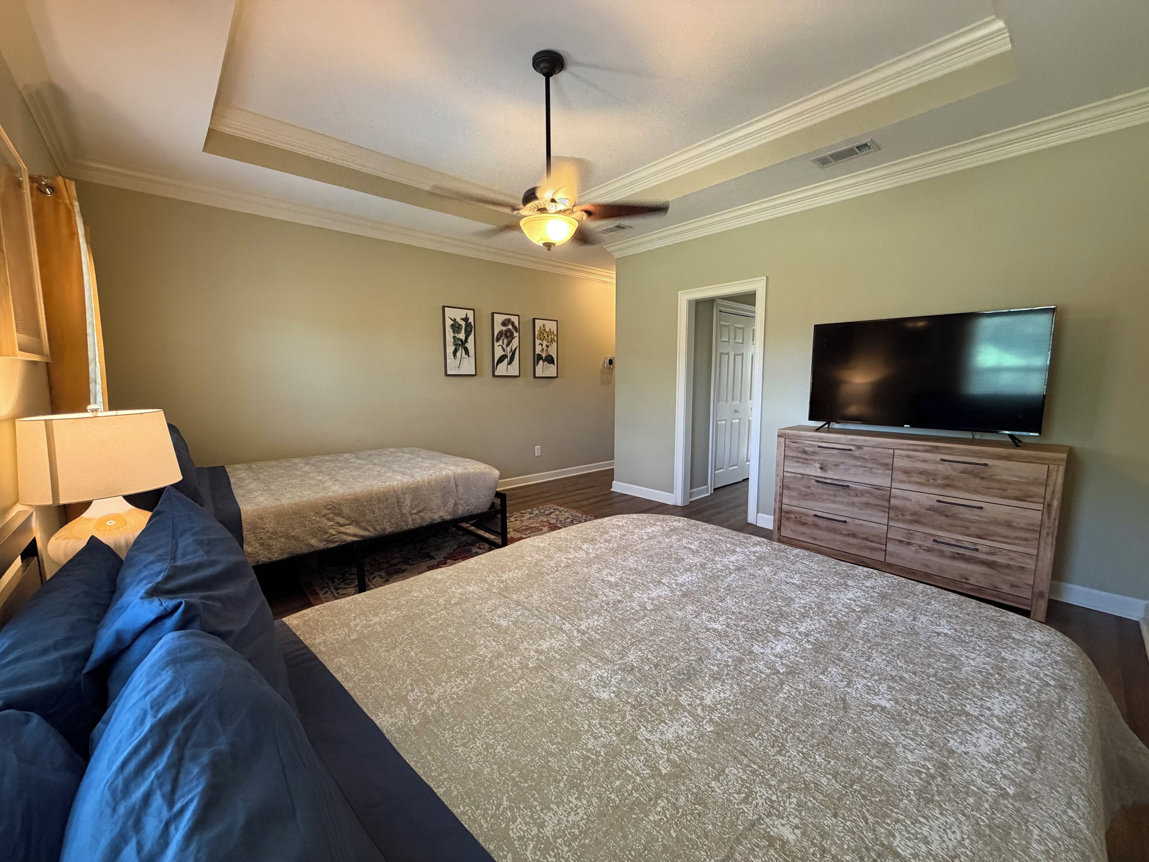 A bedroom with a large bed, a smaller bed, a dresser, a television, a ceiling fan, and framed artwork on the wall.