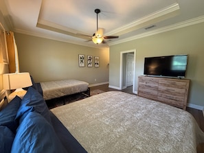 Ein Schlafzimmer mit einem großen Bett, einem kleineren Bett, einem Schminktisch, einem Fernseher, einem Deckenventilator und gerahmten Kunstwerken an der Wand.