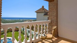 A balcony with a white balustrade, a wicker chair, and a view of a distant building with a tiled roof. - Villa Fuente by Villa Plus (Frigiliana)