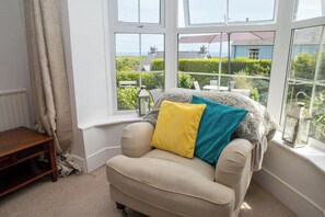 A corner room with a beige sofa, two pillows, a wooden side table, and a view of greenery and buildings through large windows. - Sea Salt, Tenby - Four Bedroom Cottage, Sleeps 8 (Penally)