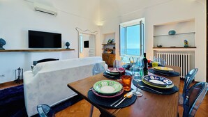 A dining area with a table set for four, a television mounted on the wall, and a view of the sea through an open door. - YourHome - La Casa di Chiara (Positano)