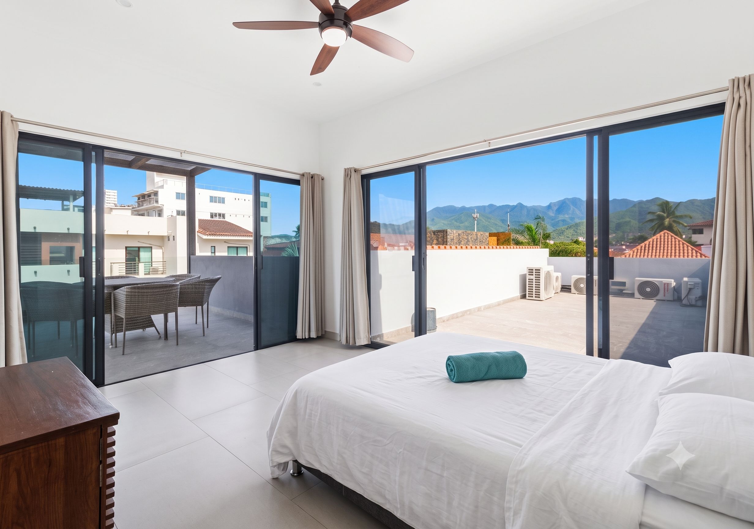 A hotel room with a bed, a ceiling fan, and a view of the city and mountains.
