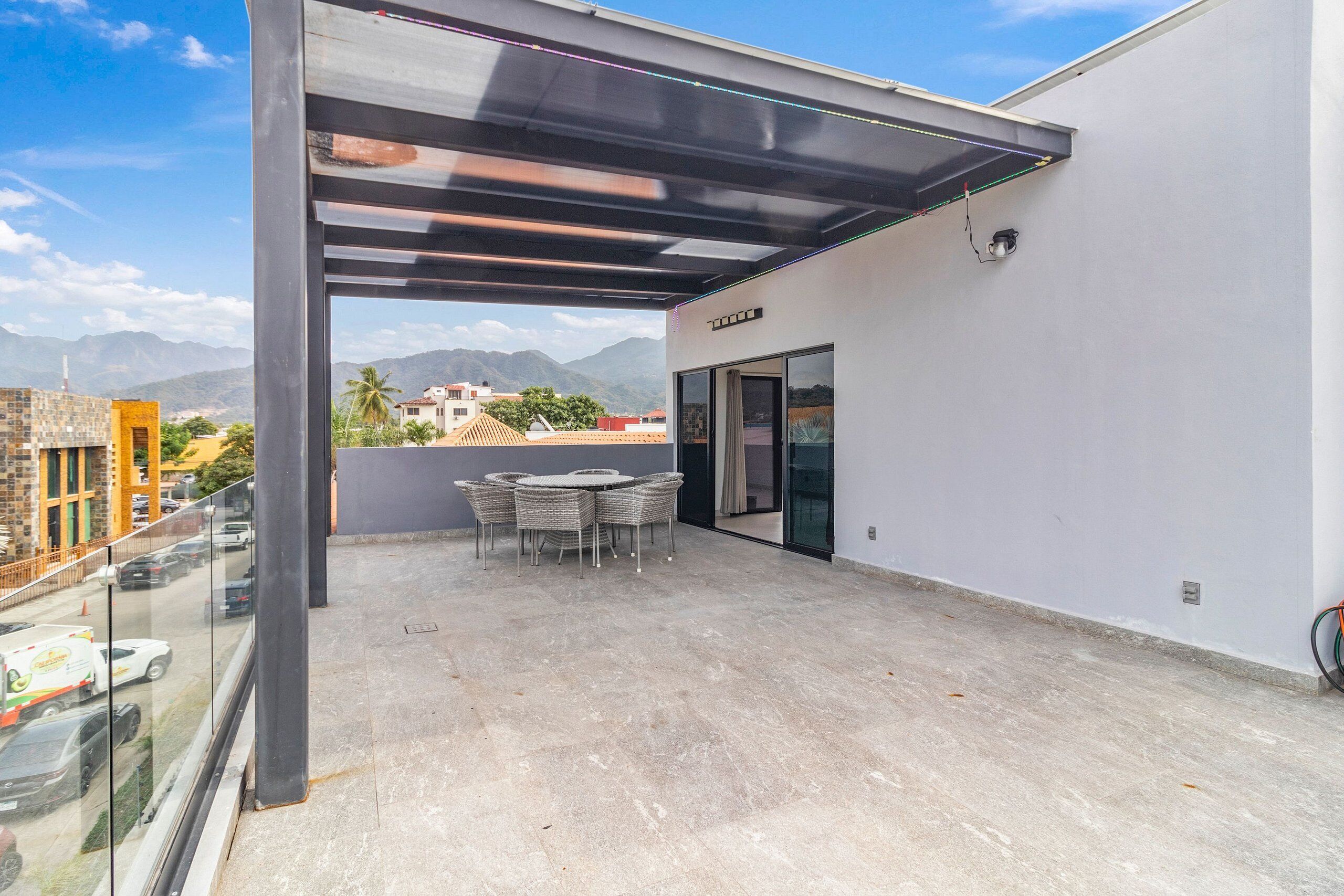 Eine Dachterrasse mit Glasbrüstung, einem Tisch mit Stühlen und Blick auf Stadt und Berge.