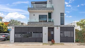 Un edificio moderno de dos pisos con entrada protegida por un portón, un balcón en el nivel superior y un pequeño jardín con flores.