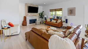 A modern living room with a stone fireplace, leather sofas, a television mounted on the wall, and a 'Pray for Surf' sign. - Charming Lakeside Escape w/ Large Deck & Firepit! (Clarkson)