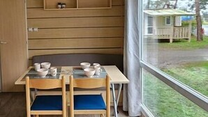 Un petit coin repas avec une table et des chaises, une fenêtre donnant sur une maison et des arbres, et un mur en bois.