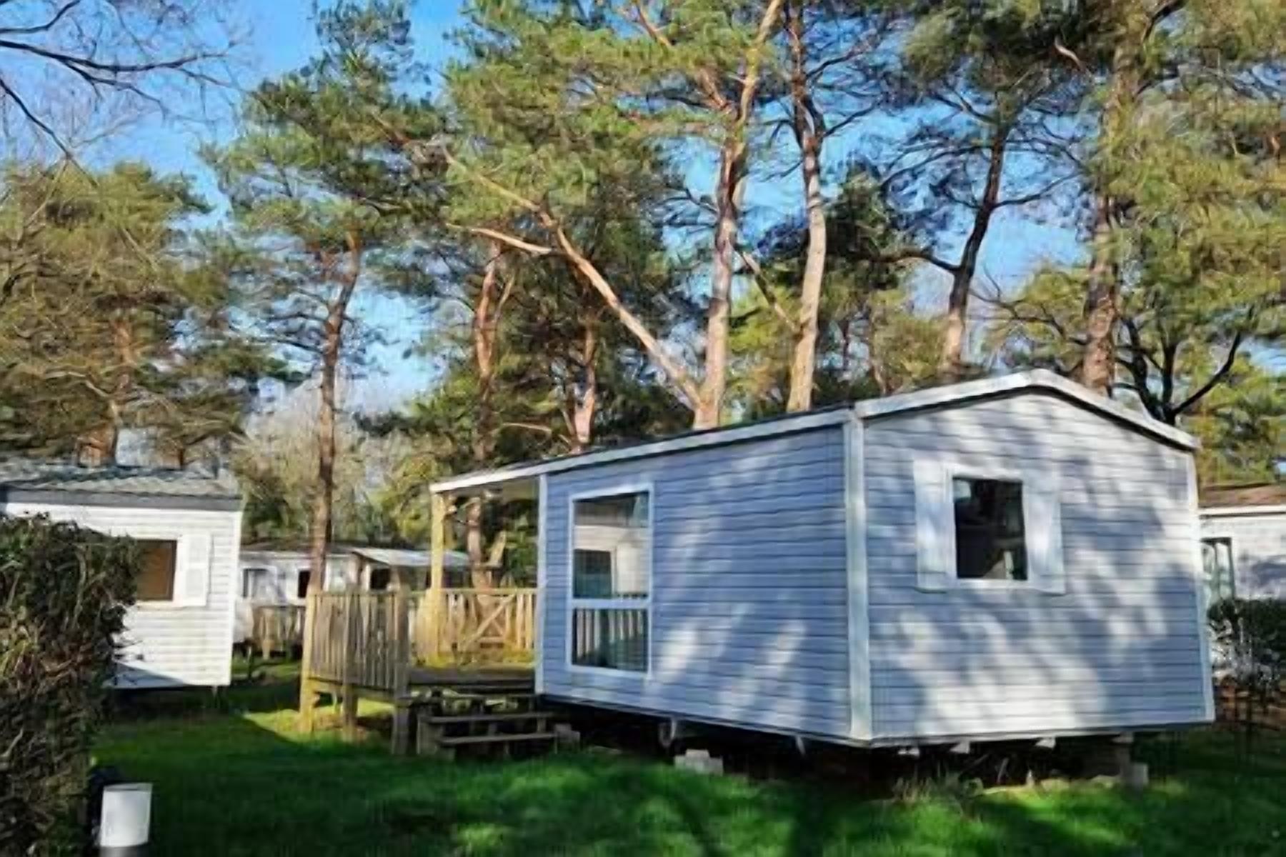 Une petite maison mobile d’un seul étage, dotée d’un porche et d’une petite terrasse, entourée d’arbres.