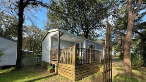 A small, single-story mobile home with a wooden deck and a porch, surrounded by trees and grass.