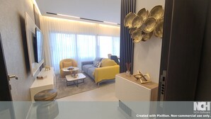 A modern hotel room with a sofa, armchair, and a coffee table. There is a large mirror, a flat-screen TV, and a decorative wall piece.
