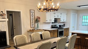 A dining area with a wooden table, four chairs, a chandelier, and a kitchen in the background. - Perfectly Pink & Cozy 2-bedroom cabin in charming Waverly Hall (Waverly Hall)