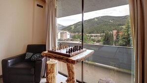 A room with a chessboard on a wooden table, a black leather chair, and a large window with a view of a forested mountain. - Pet Friendly, Spacious, Mountain Views SB102 (Copper Mountain)