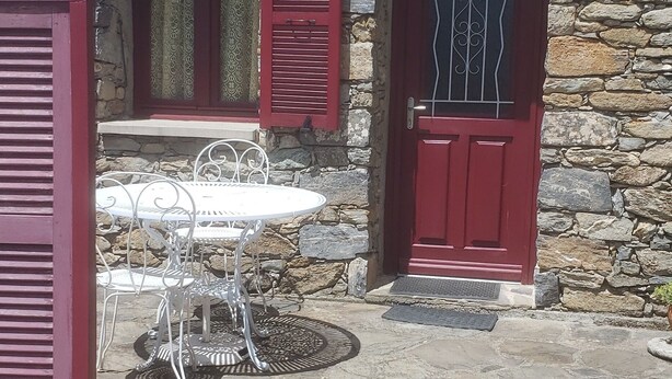 Un bâtiment en pierre avec une porte rouge et des volets aux fenêtres, une table et des chaises en métal blanc, et une cour dallée de pierres.