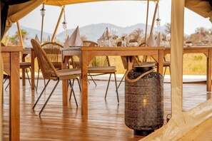 Mesa de madera con sillas y un recipiente estampado, probablemente para bebidas, en un espacio al aire libre cubierto con montañas al fondo.