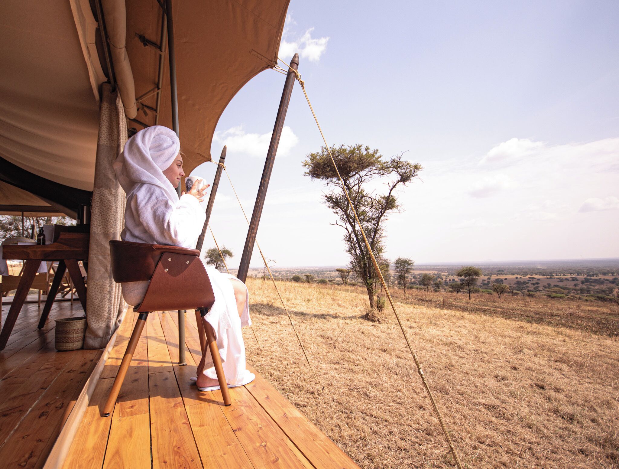 A person sitting on a wooden deck under a tent, overlooking a savannah landscape with sparse trees and a clear sky.