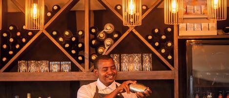 A bartender preparing a drink at a bar with wooden shelves and wine bottles in the background.