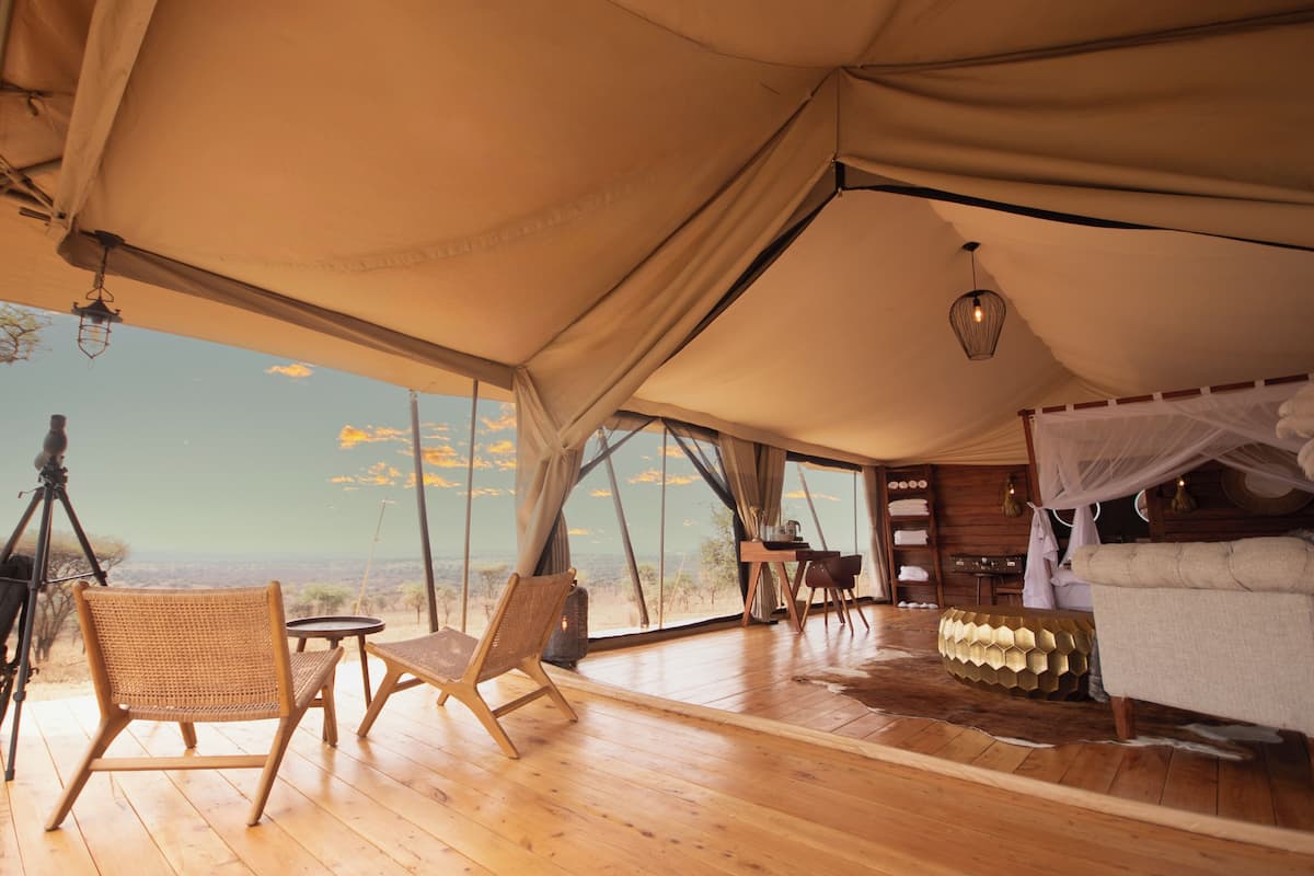 A tented room with a wooden floor, a bed with a canopy, a desk with a chair, and a view of the landscape.