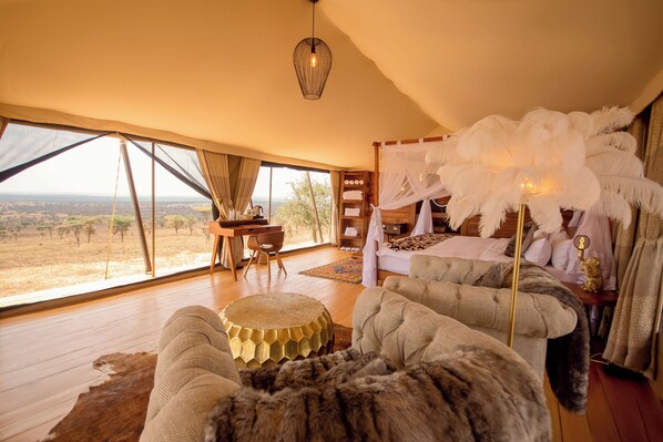Amplia habitación con un ventanal que ofrece vistas a la sabana, una cama con dosel de plumas blancas y una zona de estar acogedora con una manta de piel.
