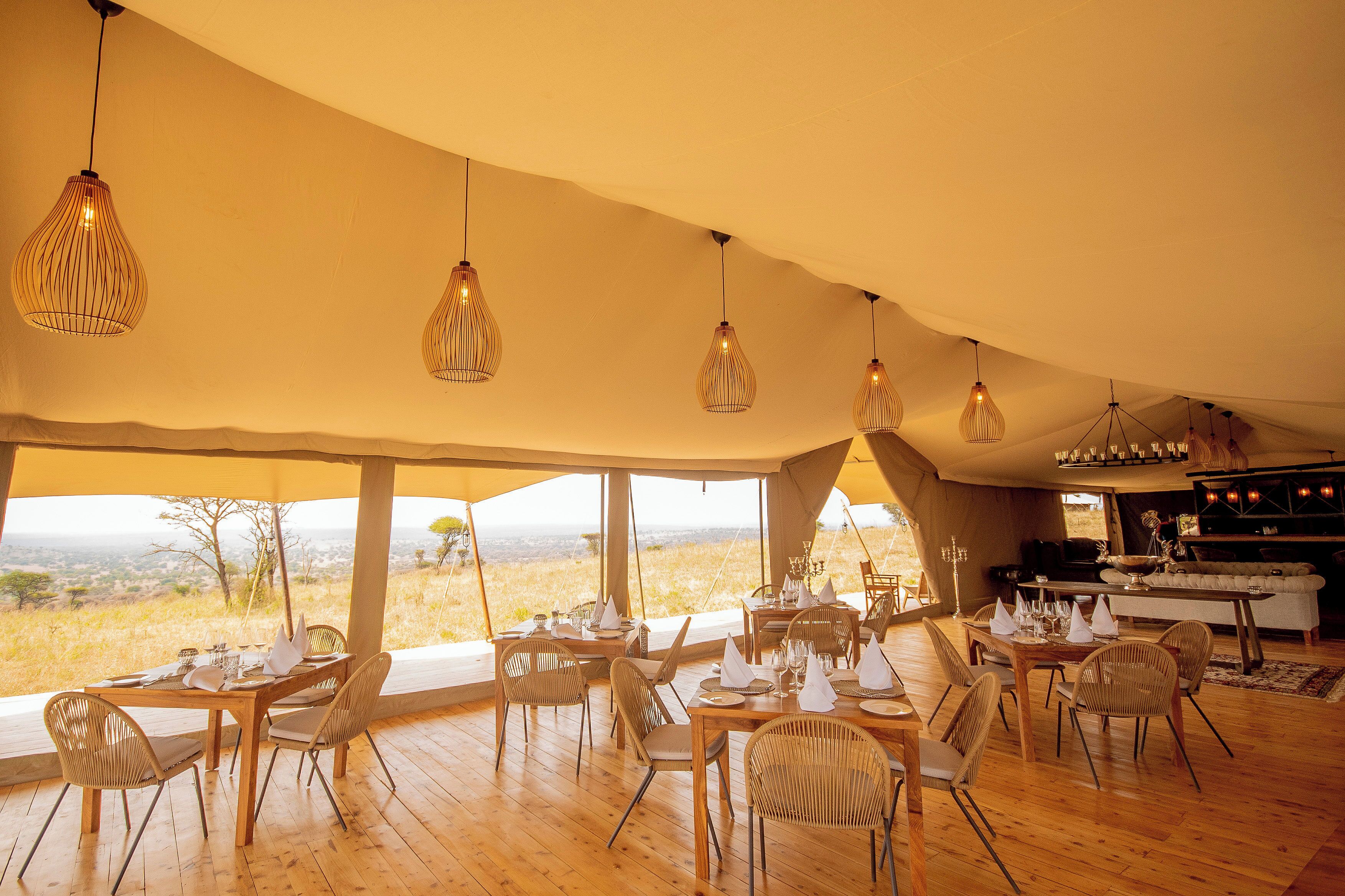 A dining area with tables set for four, large windows offering a view of the landscape, and hanging pendant lights.