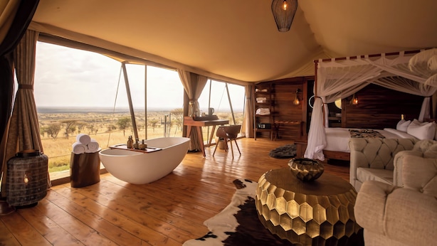 A spacious room with a large window offering a view of the savannah, a white bathtub, a wooden floor, and a cozy seating area with a patterned ottoman. - Serengeti Luxury Retreat (Serengeti)