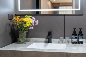 Une salle de bain avec un lavabo blanc, un vase en verre rempli de fleurs et deux bouteilles noires posées sur le comptoir.
