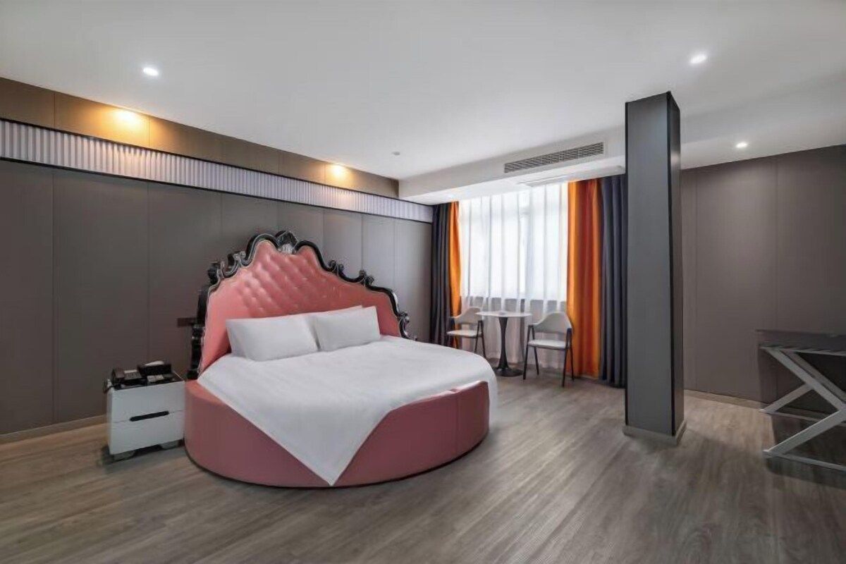 Une chambre d’hôtel moderne avec un lit rond, une petite table et des chaises, un bureau et une grande fenêtre avec des rideaux.